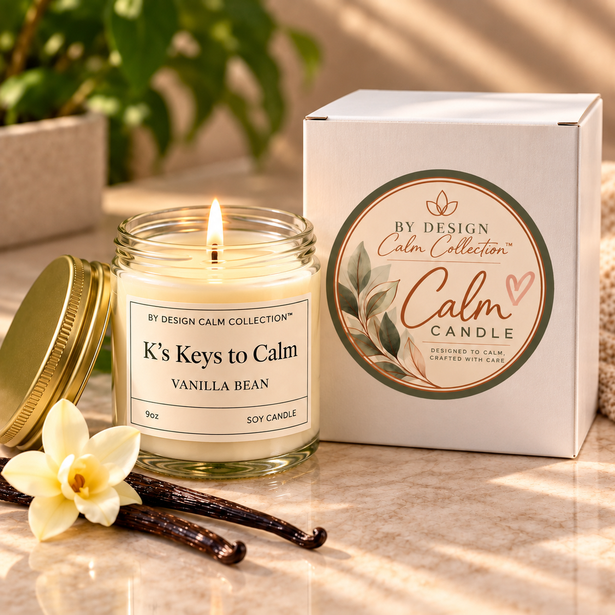 A lit glass jar candle labeled 'K's Keys to Calm Vanilla Bean' sits beside its white branded box on a marble surface, with a vanilla flower and vanilla beans in the foreground and a softly blurred indoor background.