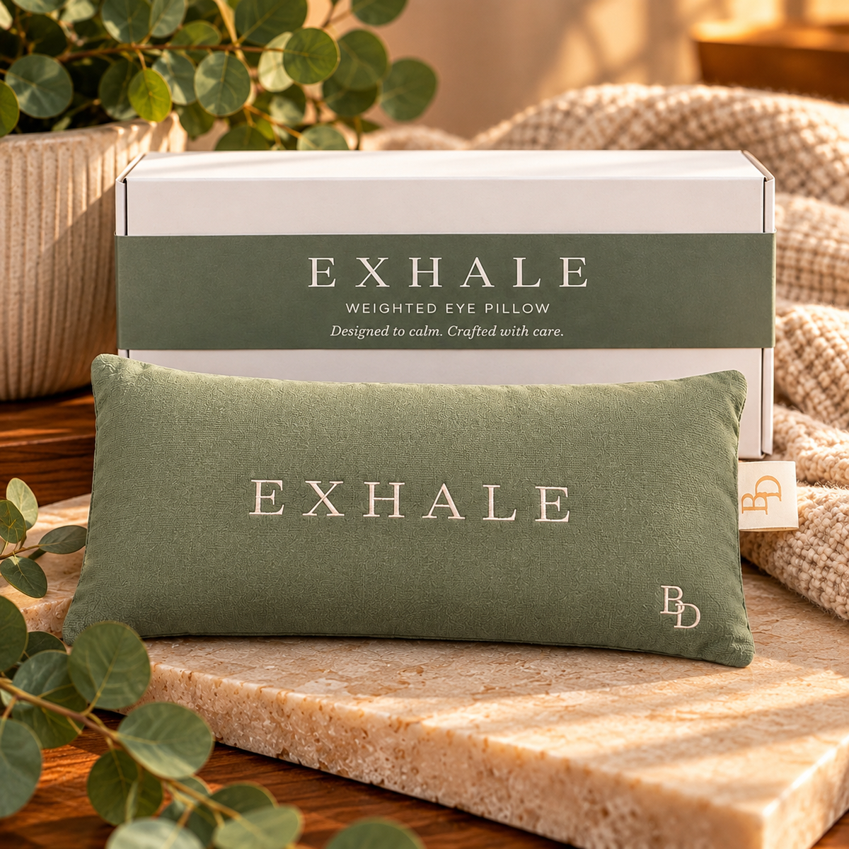 An olive green weighted eye pillow with the brand name 'EXHALE' embroidered in white on the front, displayed in front of its white and olive green packaging box. The scene is styled with a beige knit blanket, a stone slab, and eucalyptus leaves.
