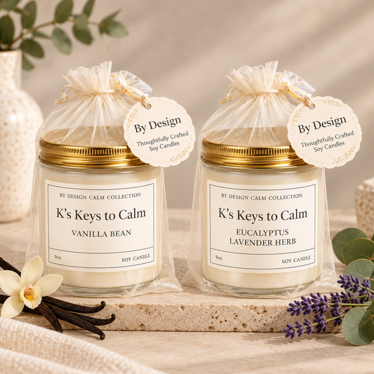 Two soy candles in clear glass jars with gold lids and cream labels, tied with sheer cream drawstring bags and hanging tags reading 'By Design.' The candles are labeled 'K's Keys to Calm' with scents 'Vanilla Bean' and 'Eucalyptus Lavender Herb.