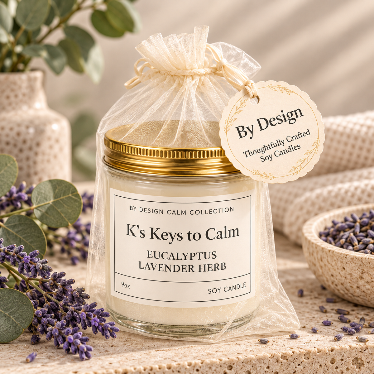A glass jar candle with a gold lid and a white label reading 'K's Keys to Calm' and 'Eucalyptus Lavender Herb,' wrapped in a sheer cream drawstring bag with a hanging tag that says 'By Design.' The candle is styled on a textured beige surface with lavender sprigs, a bowl of lavender, and soft neutral background props.