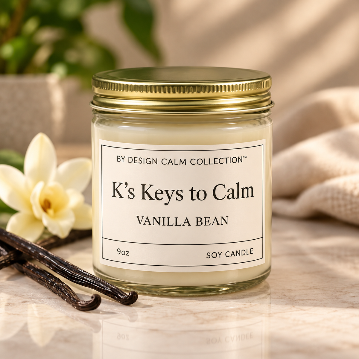 A 9oz soy candle in a clear glass jar with a gold metal lid and a cream-colored label. The label reads 'BY DESIGN CALM COLLECTION' and 'K's Keys to Calm VANILLA BEAN'.