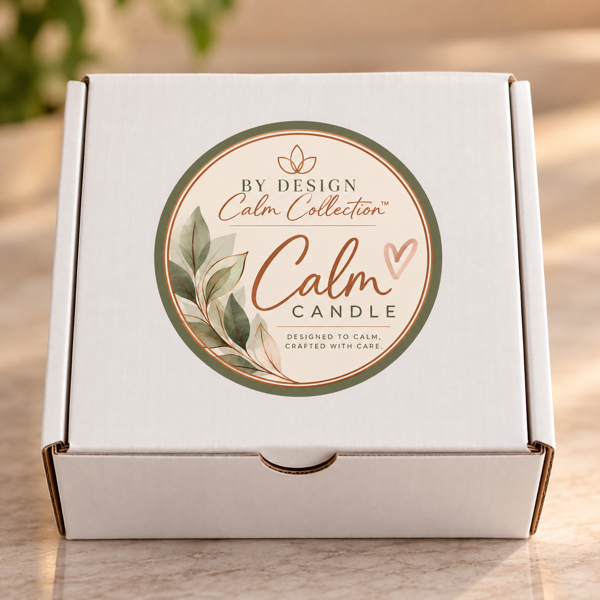 A white cardboard gift box for a 'Calm Collection' candle, featuring a circular green and beige label on the lid with the words 'By Design Calm Collection Calm Candle'.
