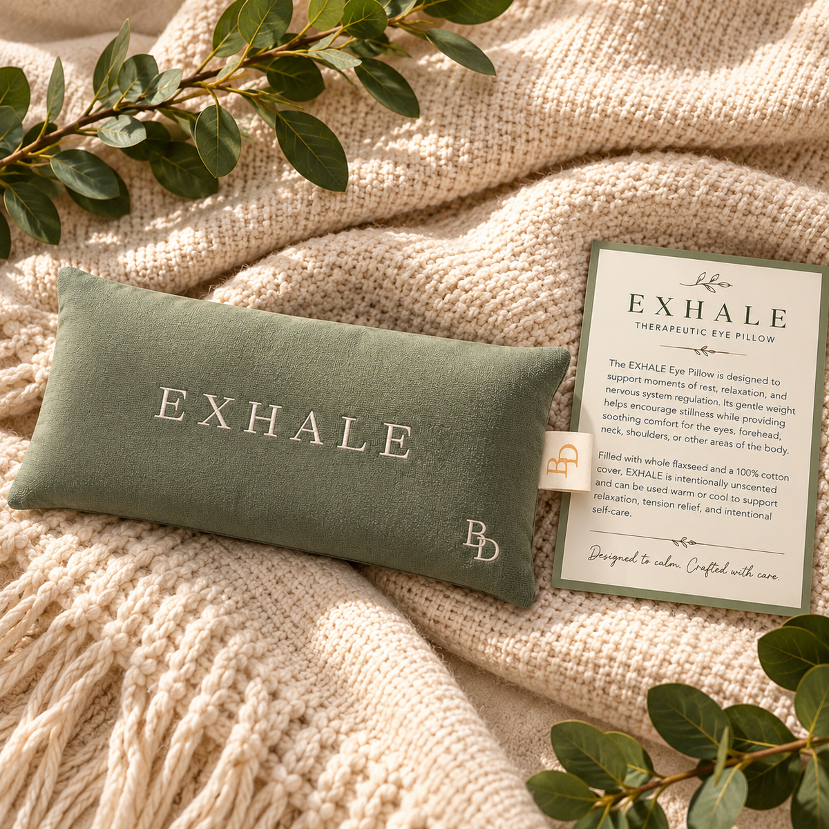 Sage EXHALE Weighted Eye Pillow styled with its therapeutic eye pillow information card on soft cream fabric.