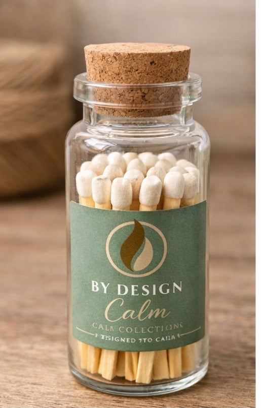 Glass bottle with cork lid containing matches labeled 'By Design Calm' on a wooden surface.