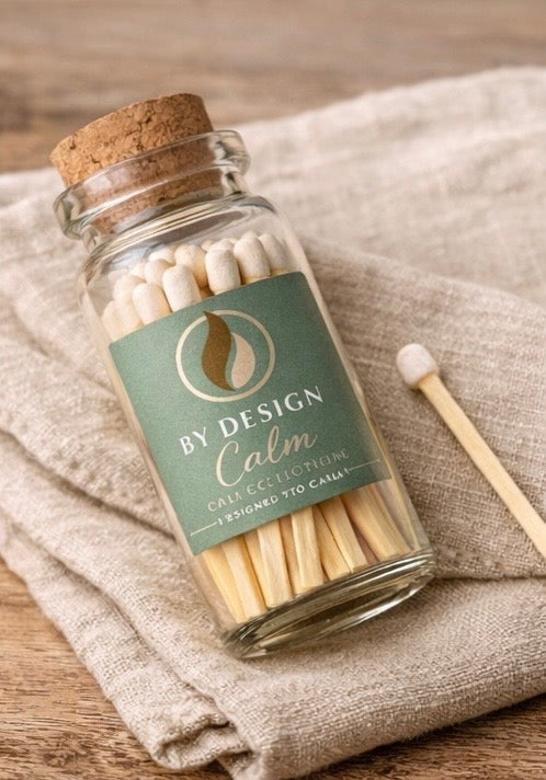 Jar of matches with a 'By Design Calm' label on a textured fabric background