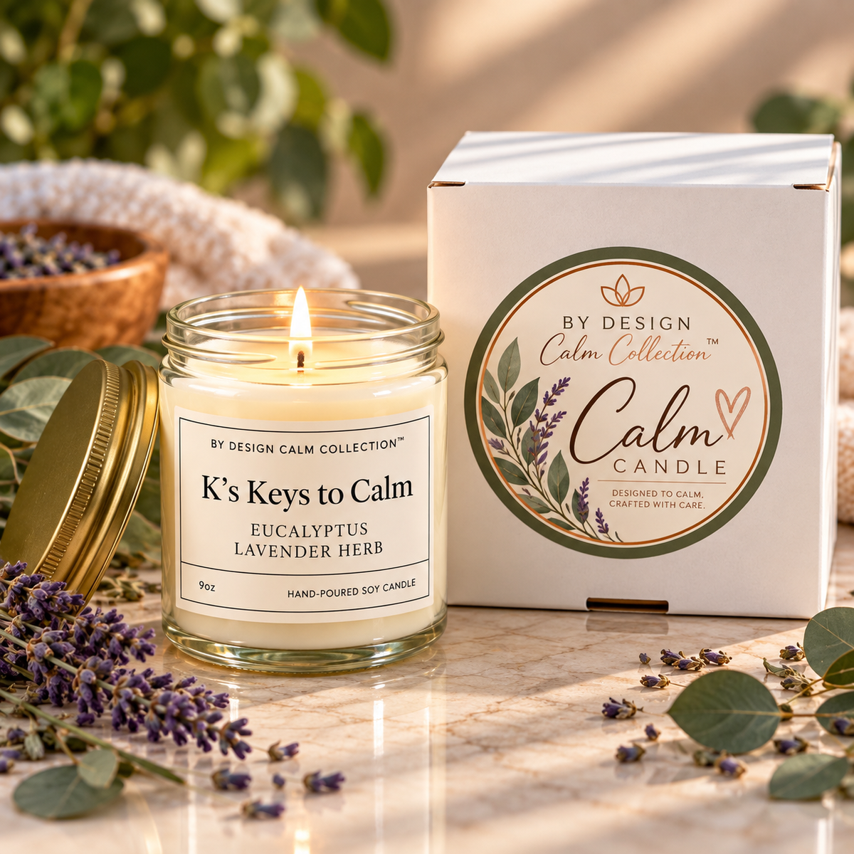 A lit glass jar candle labeled 'K's Keys to Calm' with a gold lid beside its white product box, styled on a marble surface with lavender sprigs, eucalyptus leaves, and soft natural background elements.