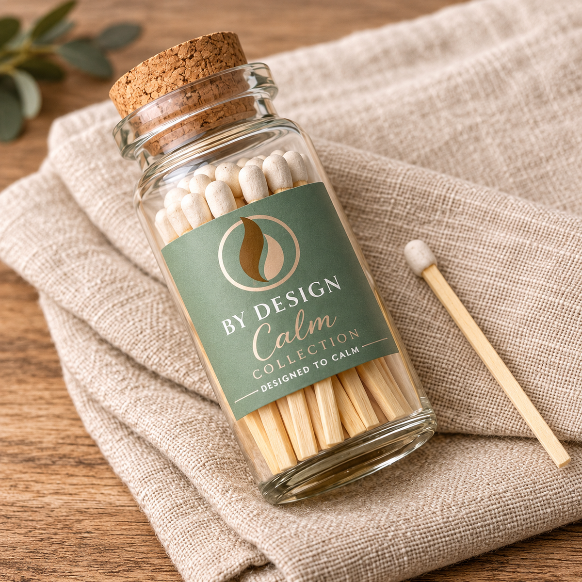 A clear glass jar with a cork stopper and a green label reading 'BY DESIGN Calm COLLECTION' sits on a beige linen cloth over a wooden surface. The jar contains wooden matches, and one match lies beside it.